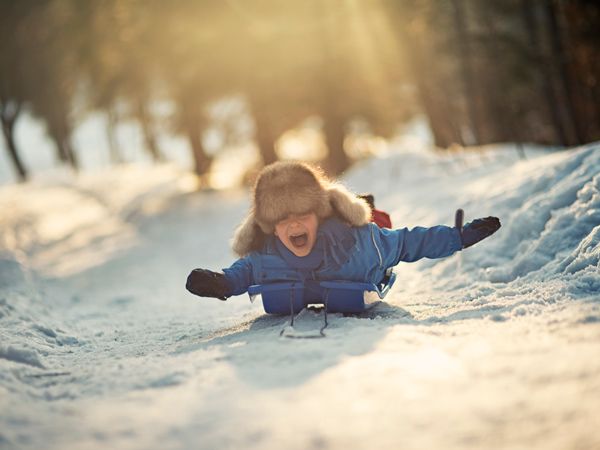 Little boy in funny fake fur hat sliding on his sled and yelling. Sunny winter day evening.