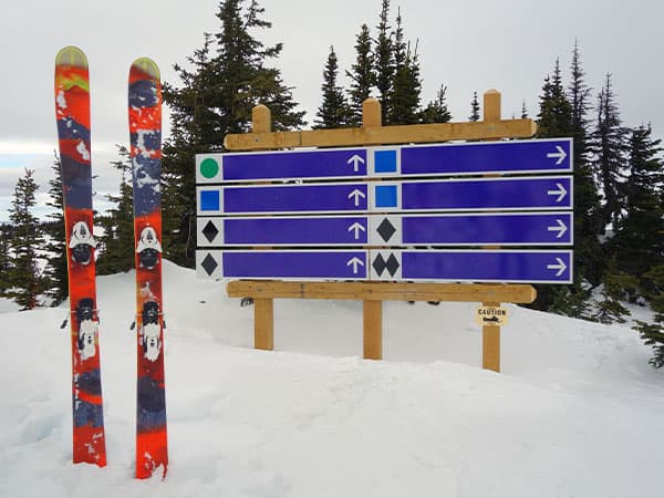 Pair of skis sitting upright in a snow bank at the top of a chairlift next to a wooden ski-run sign. Taken at Sun Peaks Resort, British Columbia, Canada