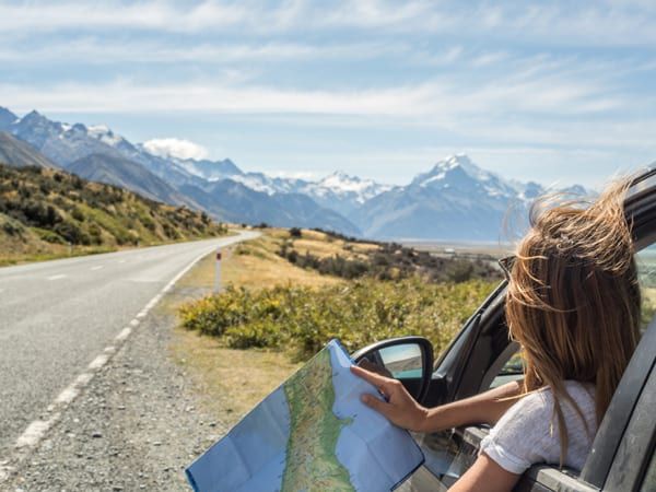 A woman in a car hangs out the window looking at a map, with snow-capped mountains in the distance.