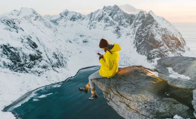 Woman sitting on cliff and using smartphone on Lofoten island in snow.