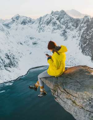 Woman sitting on cliff and using smartphone on Lofoten island in snow.