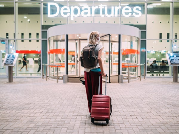 A woman stands at the departures entrance at the airport with a suitcase and a backpack.