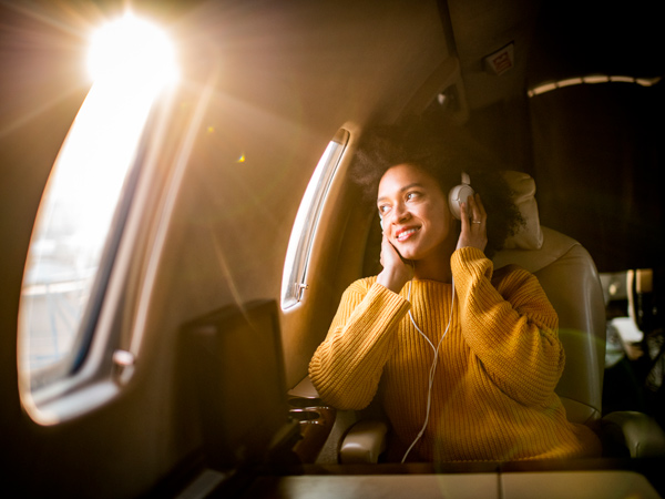 Young modern woman sitting in a private jet, listening to music through the headphones and looking through the window