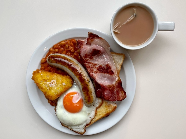 a plate of sausage, eggs, bacon and hash brown with coffee at Norman’s Cafe, London
