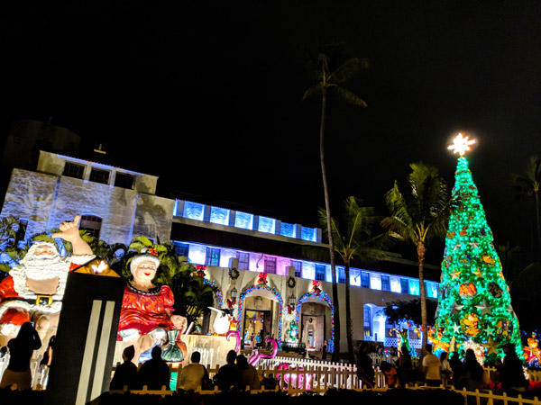  People look at Mr. and Mrs. Claus sit on top waterfall with Turtle figure holding tray with coconut drink, poi bag, and Lau lau Light up and Christmas Tree with star at night in front of Honolulu Hale, the Mayor Office, as part of Honolulu City Lights