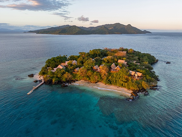 An aerial view of Royal Davui Island Resort reveals a secluded paradise. Lush palms, thatched villas, and white sands surrounded by crystal-clear turquoise waters.