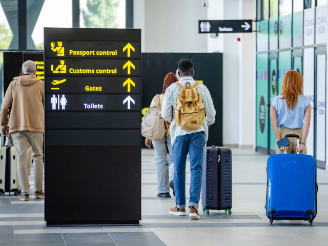 Groups of individuals walk towards passport control and customs at an airport, with signs showing directions, illustrating travel and passage through security processes.