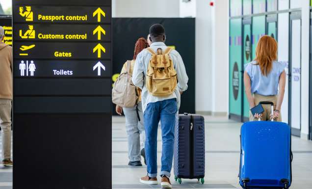 Groups of individuals walk towards passport control and customs at an airport, with signs showing directions, illustrating travel and passage through security processes.