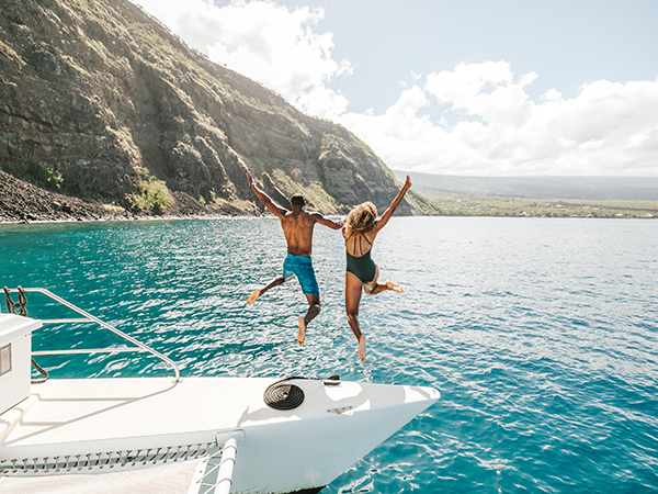 Two people jumping off a yacht with the beautiful coastline of Hawai‘i in the background.