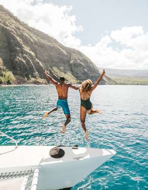 Two people jumping off a yacht with the beautiful coastline of Hawai‘i in the background.