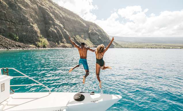 Two people jumping off a yacht with the beautiful coastline of Hawai‘i in the background.