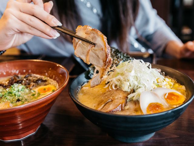 Woman hand pinching noodle in Ramen Pork Bone Soup (Tonkotsu Ramen) with Chashu Pork, Scallion, Sprout, Corn, Dried Seaweed and boiled egg served in black bowl